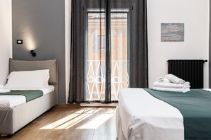 A hotel room with two beds, a view of the outside, a painting on the wall, and a black radiator. - Saragozza Apartments (Bologna)