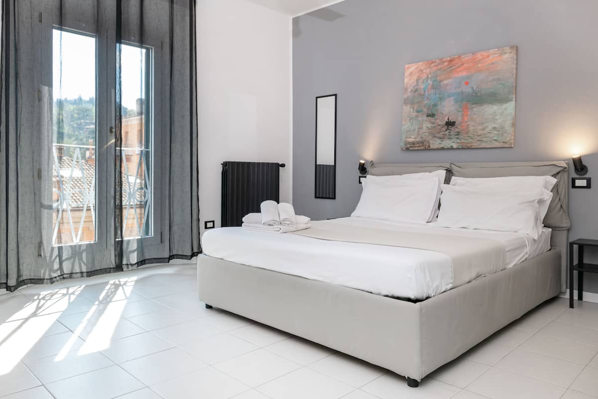 A modern bedroom with a large bed, a painting on the wall, and a view through the window.