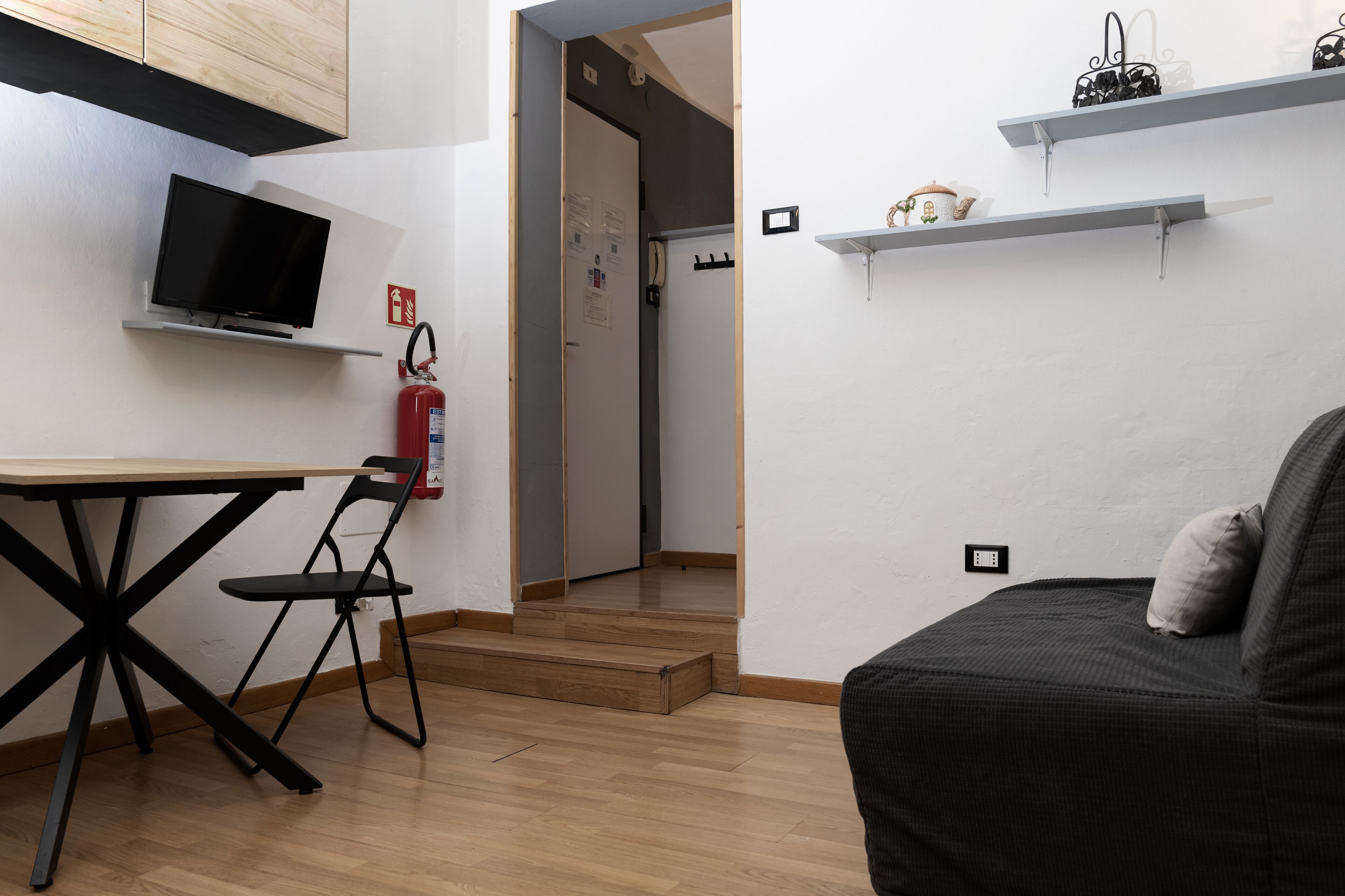 Una habitación pequeña y moderna con escritorio, silla y sofá negro. Hay un televisor montado en la pared, un extintor y una puerta que conduce a otra habitación.