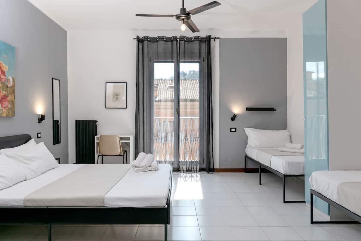 A hotel room with a bed, two smaller beds, a chair, a desk, a ceiling fan, and a window with curtains.