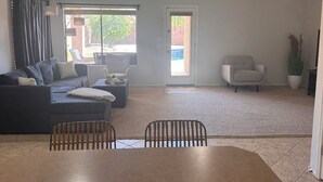 A spacious living room with a dark-colored sofa, a dining table with chairs, a ceiling fan, and a large window with curtains.