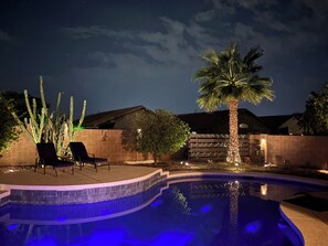 A swimming pool with a curved edge, surrounded by a brick wall. There are two lounge chairs and a palm tree with lights.