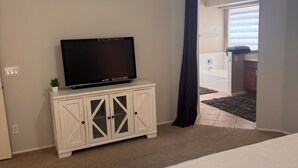 A bedroom with a bed, a TV on a white cabinet, a painting on the wall, and a view of a bathroom through an archway.