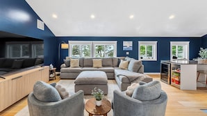 Een moderne woonkamer met een hoekbank, een poef en grijze fauteuils. Er is een houten salontafel met een vaas, een televisie en een klein bijzettafeltje met een lamp.