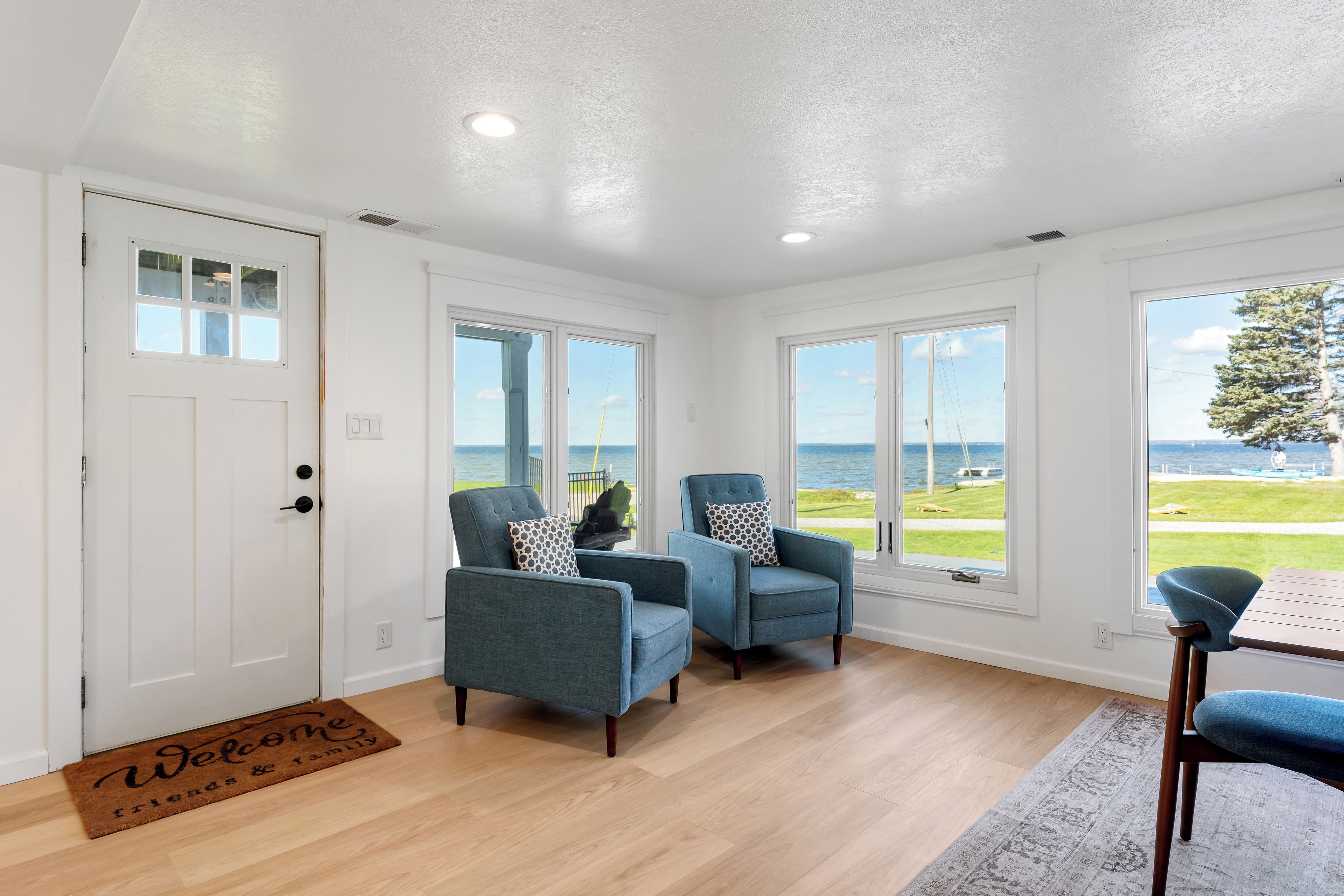 A room with a wooden floor, two blue armchairs, a small table, and a view of the sea through large windows.