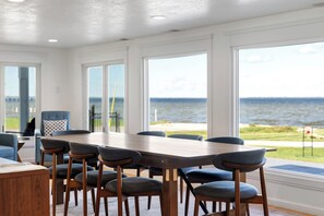 A dining area with a large table and chairs, large windows with a view of the sea, and a view of the outdoors. - Large 5BD Lakehouse w/ Beach & Hot Tub (Prudenville)