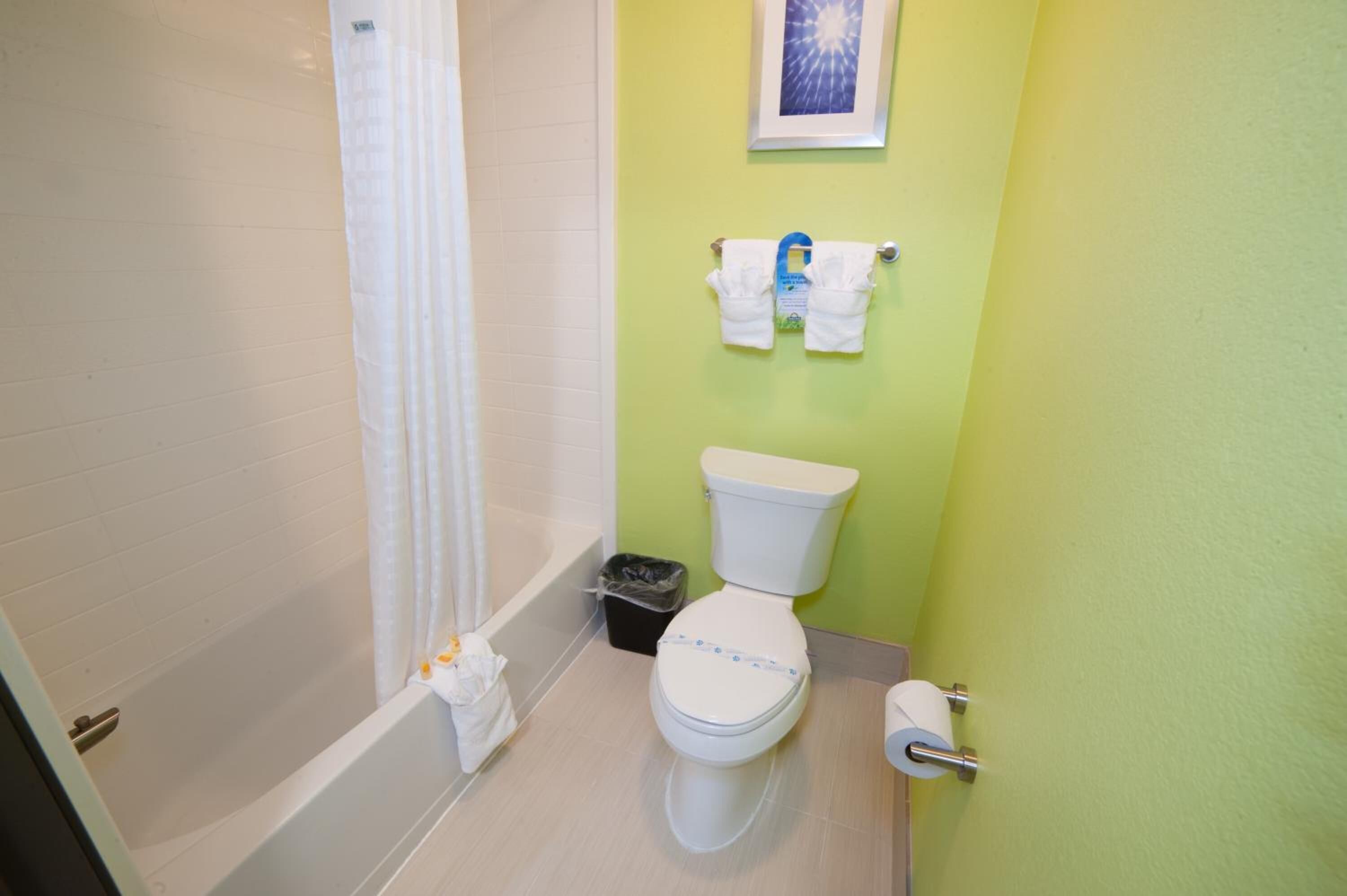 A bathroom with a toilet, a bathtub, a shower curtain, a trash bin, and a towel rack with towels.