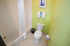 A bathroom with a toilet, a bathtub, a shower curtain, a trash bin, and a towel rack with towels. - Arya Inn & Suites Farmers Branch (Farmers Branch)