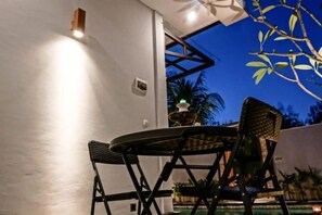 An outdoor seating area with a table and chairs, a white wall, and a view of a pool and palm trees. - Muaya Villa (Bali)