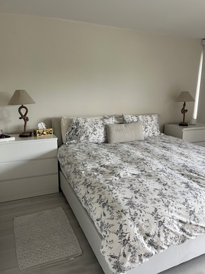 Une chambre à coucher avec une couvre-lit à motifs floraux, deux tables de chevet avec des lampes, une boîte à bijoux et un petit tapis.