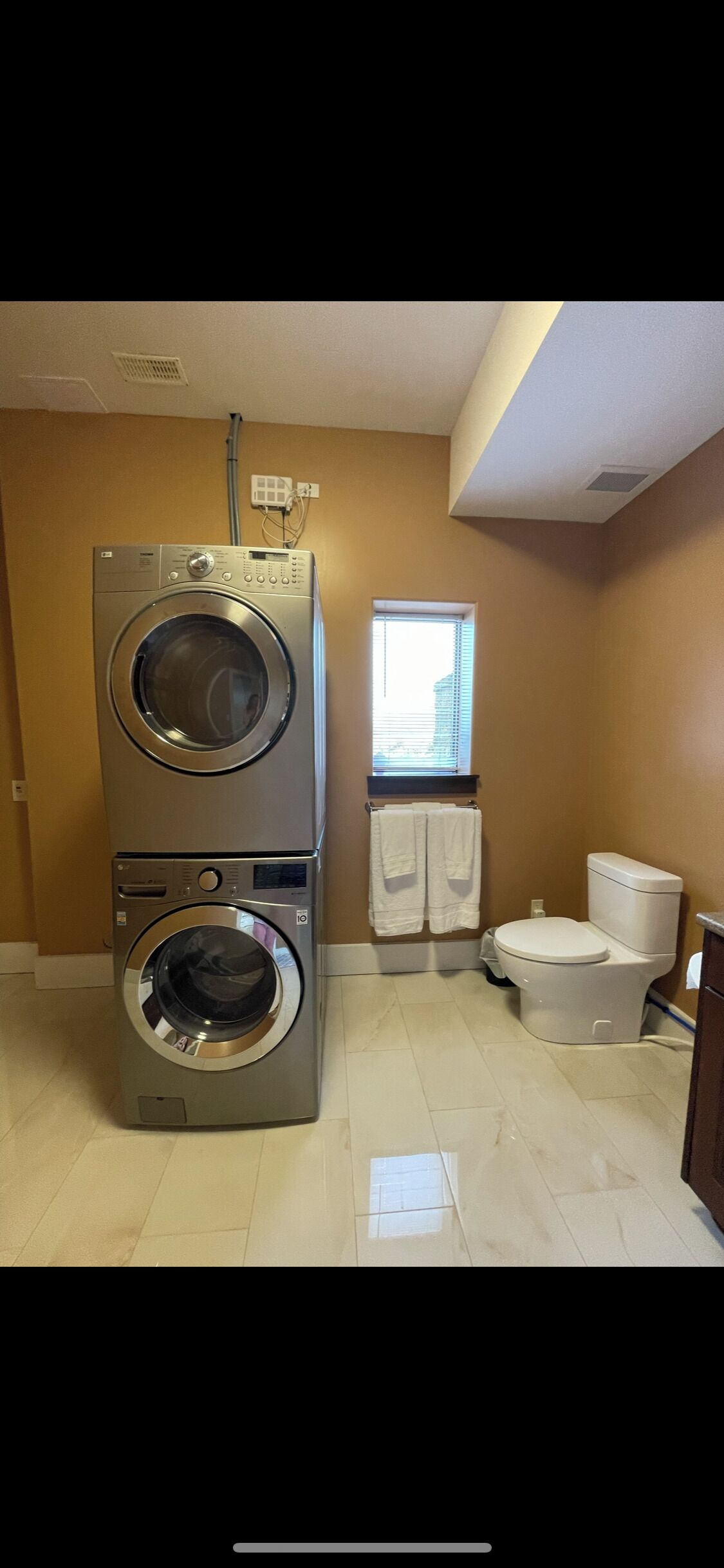 A laundry room with stacked washers and dryers, a toilet, and a window with blinds.
