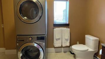A laundry room with stacked washers and dryers, a toilet, and a window with blinds.