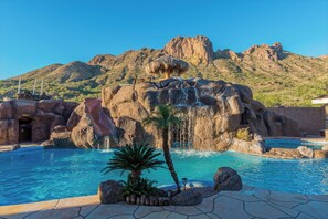A swimming pool with a waterfall feature, surrounded by rocks and palm trees, with a mountainous backdrop.