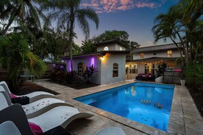 Une piscine avec des chaises longues, un petit bâtiment dont l’extérieur est illuminé de lumière violette, et des palmiers.