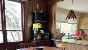 A wooden dining room with a round table and chairs, a kitchen area with wooden cabinets, and a large window providing natural light. - Rustic Cabin Getaway Near Kenora (Kenora)