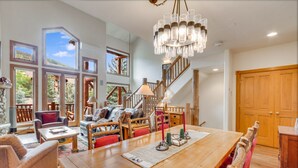 A spacious living area with a wooden dining table, comfortable seating, a staircase, and large windows offering a view of the outdoors. - Ski Shuttle - Steps to Pool & Hot Tub - Covered Parking (Steamboat Springs)