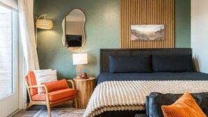 A bedroom with a bed, a chair, a nightstand, a lamp, a mirror, and a wall-mounted picture.