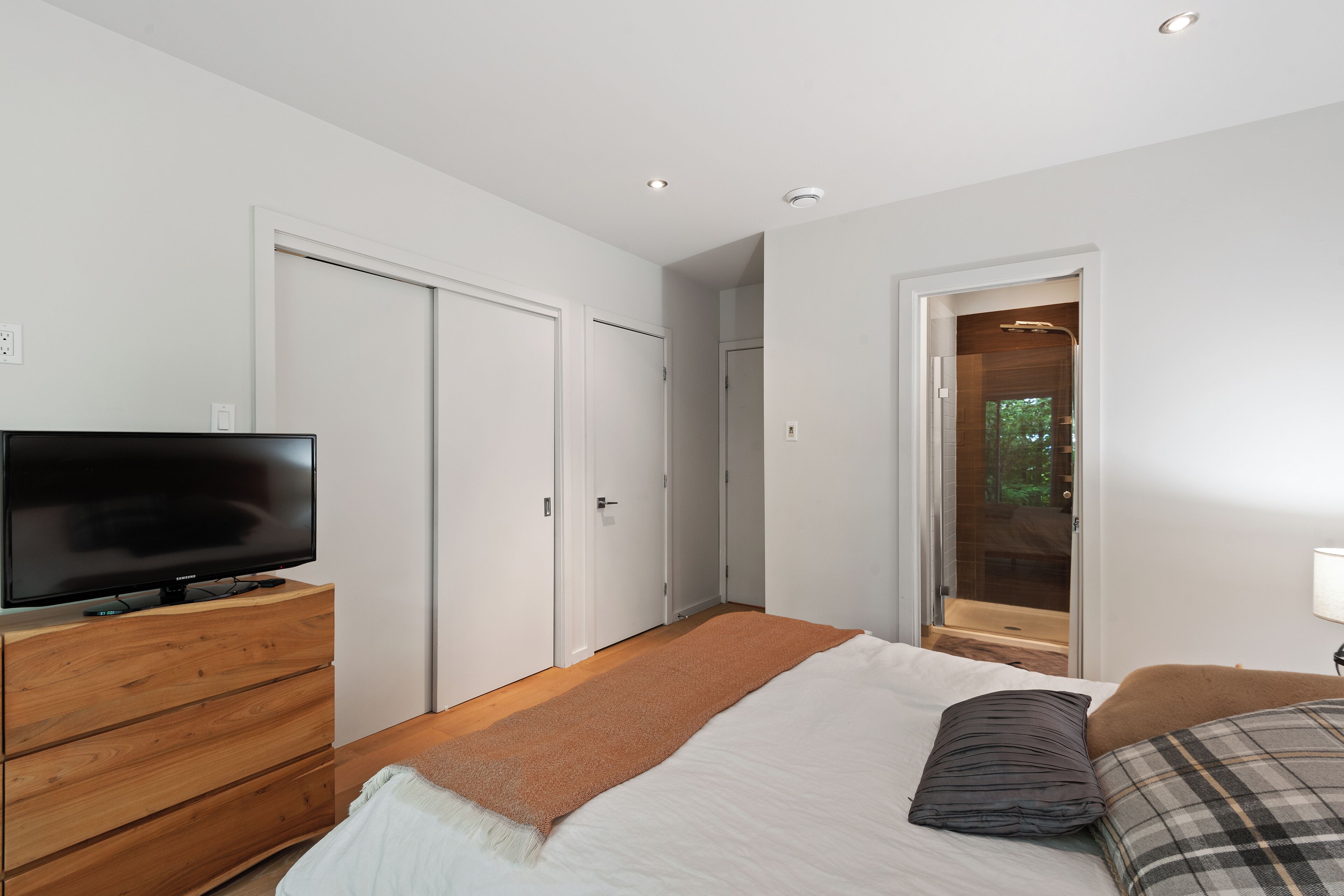 A bedroom with a bed, a TV on a wooden dresser, a sliding door leading to a shower area, and a view of greenery outside.