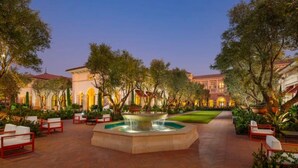 Un patio al aire libre con una fuente, zonas de estar y un edificio con ventanas arqueadas al fondo.