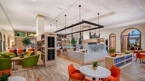 Un lobby moderno de hotel con asientos verdes y anaranjados, un bar central y una vista a través de un arco.