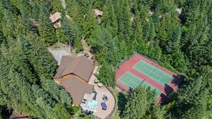 Vue aérienne d’une propriété comprenant un court de tennis, une piscine et un bâtiment entouré d’une forêt dense.