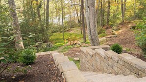 A stone staircase leading up a hill in a forested area with a wooden bridge and a stone wall.