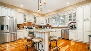 A spacious kitchen with white cabinets, a central island with stools, stainless steel appliances, and a wooden floor.