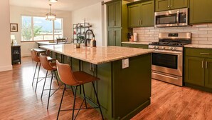Une cuisine avec des meubles de couleur verte, un grand îlot central doté de tabourets, des appareils électroménagers en acier inoxydable et une vue sur l’extérieur grâce à une fenêtre.