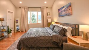 Une chambre à coucher avec un grand lit, une table de chevet, une chaise, une commode, une lampe, un tableau, une fenêtre avec des rideaux, une plante et une étagère avec des livres et un vase.