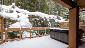 A wooden deck with a hot tub, surrounded by snow-covered trees and a rocky cliff. - Northern Lights 32 | Ski-in/out Luxury Townhome, Hot Tub, (Whistler)