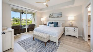A bedroom with a large bed, two bedside tables with lamps, a ceiling fan, a sliding glass door to a balcony, and a view of palm trees. - Charming Three Bedroom Villa in Beautiful Naples (Naples)