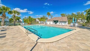 A rectangular swimming pool with a tiled deck, surrounded by lounge chairs and umbrellas, with palm trees and a clear blue sky in the background. - Quiet, Turn-Key, Newly Furnished, Beautiful! CLOSE to EVERYTHING! (Fort Myers)