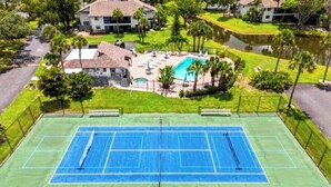 A tennis court surrounded by a fence, with a pool and lounge chairs nearby. - Quiet, Turn-Key, Newly Furnished, Beautiful! CLOSE to EVERYTHING! (Fort Myers)