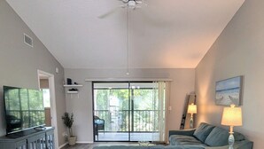Una sala de estar con un sofá, una televisión, un ventilador de techo y un gran ventanal corredizo que da a un balcón.
