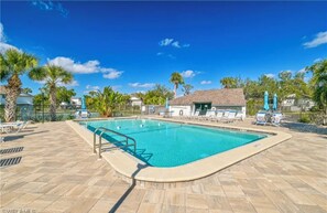 A rectangular swimming pool with a tiled deck, surrounded by lounge chairs and umbrellas, with palm trees and a clear blue sky in the background.