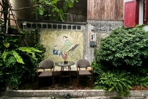 A small outdoor seating area with a table and two chairs, surrounded by lush greenery and a decorative wall with a mural. - Photohostel (Bangkok)