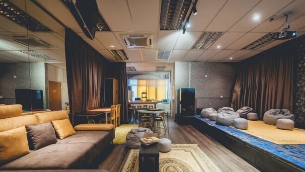 A modern living room with a sofa, a coffee table, and bean bags. There is a dining area in the background with a table and chairs. - Tun Aminah Shoplot 10-14pax KTV Mahjong Funhouse (Sekudai)