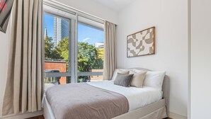 Une chambre dotée d’un grand lit, d’une fenêtre avec des rideaux, d’une vue sur la ville et d’une œuvre d’art abstraite encadrée accrochée au mur.