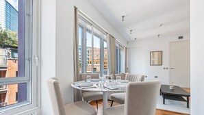 Une salle à manger moderne avec une table ronde dressée pour quatre personnes, de grandes fenêtres et une vue sur les bâtiments environnants.