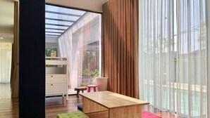 Ein modernes Zimmer mit Holztisch, bunten Kissen, Blick durch leichte Vorhänge und einer Schiebeglastür.