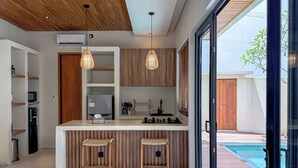 A modern kitchen with wooden cabinets, a central island with stools, and a view of a swimming pool. - Athaya Canggu Villas by Nagisa Bali (Canggu)