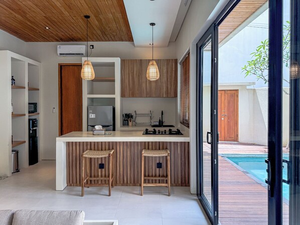 A modern kitchen with wooden cabinets, a central island with stools, and a view of a swimming pool. - Athaya Canggu Villas by Nagisa Bali (Canggu)
