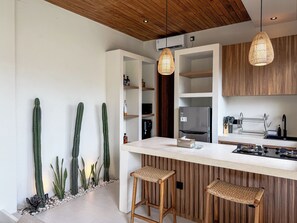 A modern kitchen with a wooden ceiling, white walls, and a central island with bar stools. There are two hanging pendant lights, a refrigerator, and a sink with a knife block. - Athaya Canggu Villas by Nagisa Bali (Canggu)