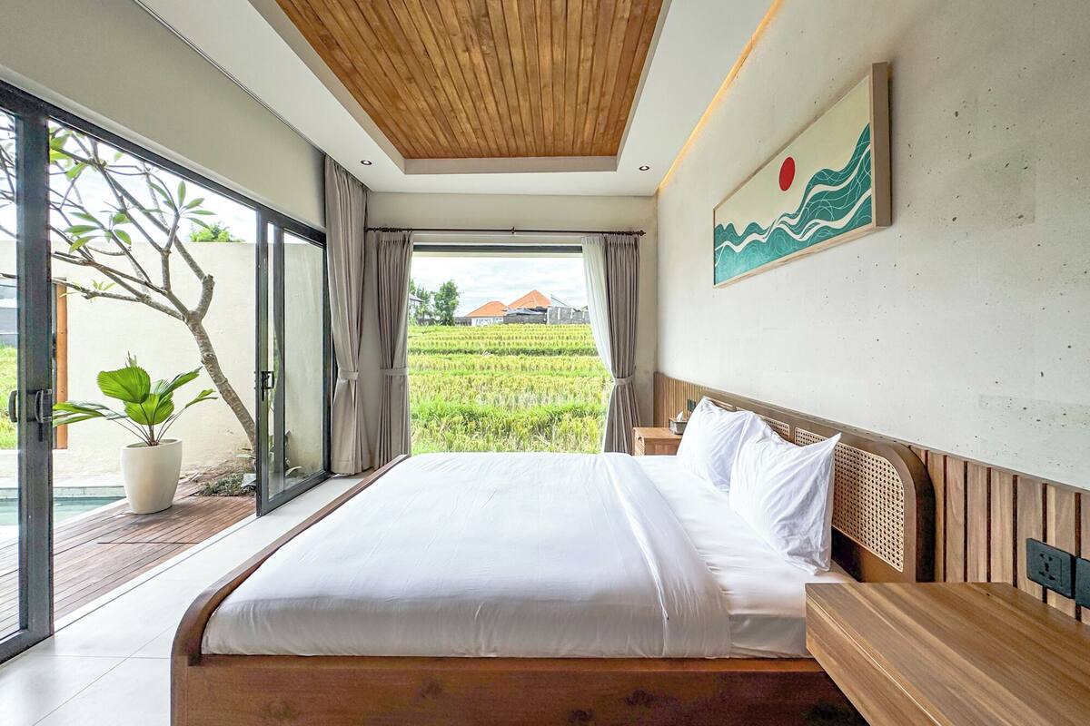 A modern bedroom with a large bed, wooden headboard, and a view of a rice field.