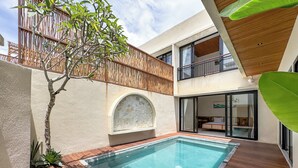 A modern pool area with a wooden deck, a stone wall, and a glass door leading to a bedroom. - Athaya Canggu Villas by Nagisa Bali (Canggu)