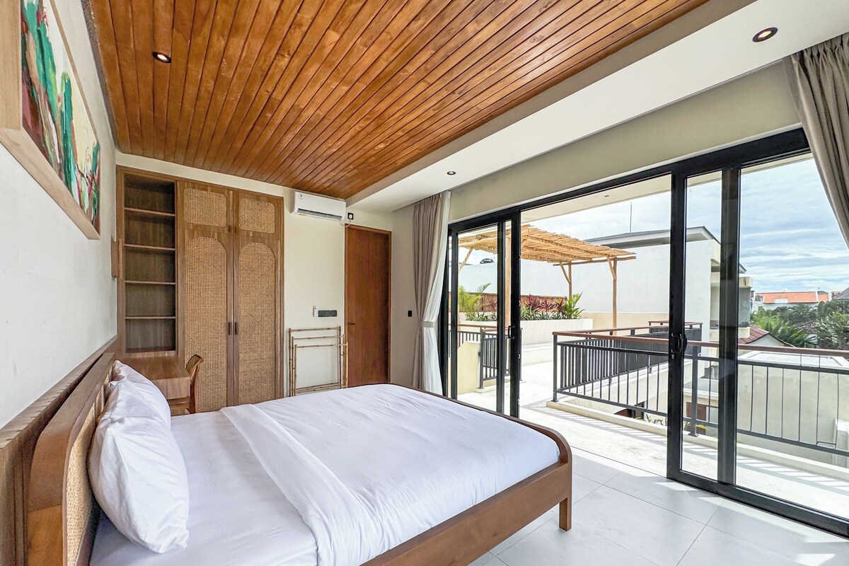 A bedroom with a wooden ceiling, a large bed, a wardrobe, and a sliding glass door leading to a balcony with a view of the sea.