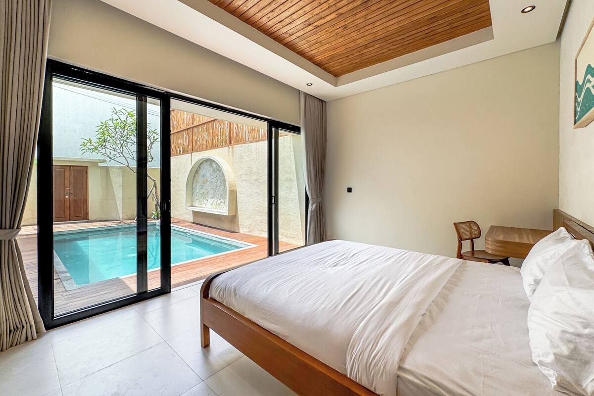 A bedroom with a large bed, a view of an outdoor swimming pool, and a wooden ceiling.