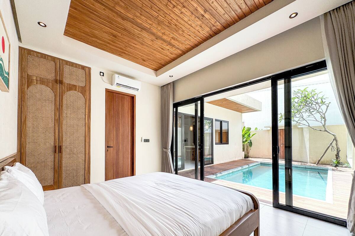 A bedroom with a large bed, wooden ceiling, and a sliding glass door leading to a swimming pool.