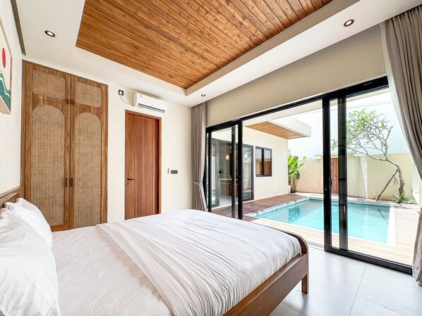 A bedroom with a large bed, wooden ceiling, and a sliding glass door leading to a swimming pool. - Athaya Canggu Villas by Nagisa Bali (Canggu)