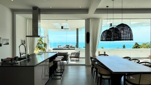 A modern kitchen with a large island, bar stools, and a dining area with a long table and chairs. - Golden Lotus Villas (Koh Samui)